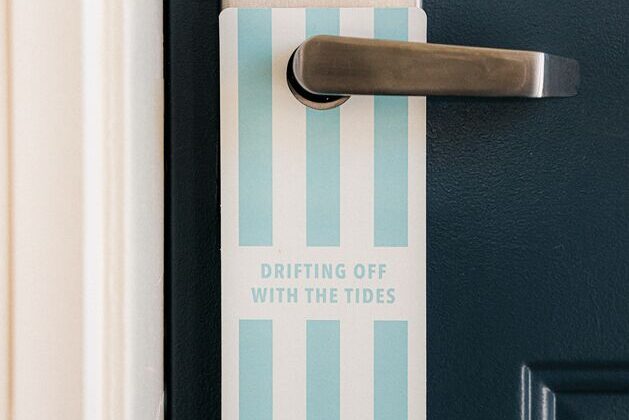 A blue and white striped door hanger with the text DRIFTING OFF WITH THE TIDES hangs on a silver door handle attached to a dark blue door.
