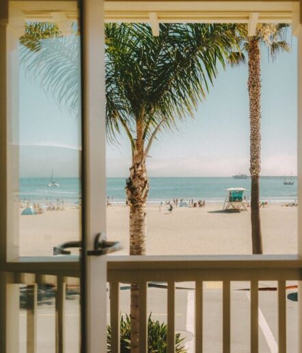 View of a sandy beach with palm trees, people, and sailboats on the ocean, seen through a glass door and a white balcony railing.