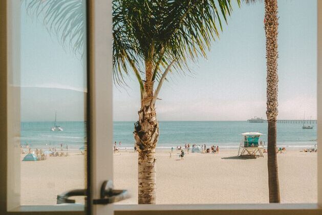View through a window of a sunny beach with palm trees, people relaxing on the sand, sailboats on the ocean, and a distant pier under a clear sky.