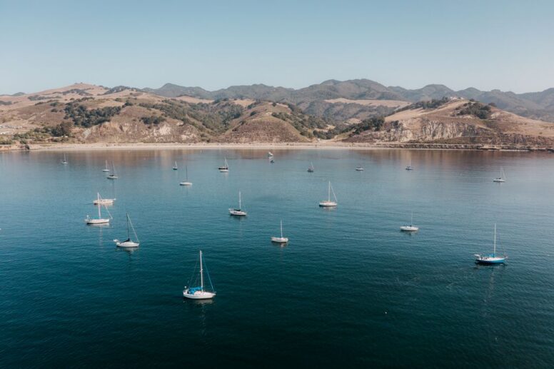 Sailboats are anchored in calm blue water near a coastline with rolling hills and mountains in the background under a clear sky.