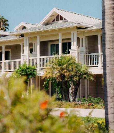 A two-story beige house with white trim features a large front porch and balcony, surrounded by palm trees and greenery under a clear blue sky.
