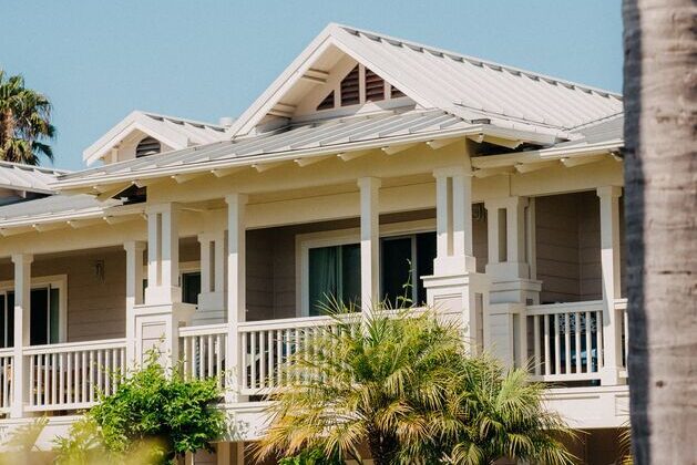 A beige house with white trim features a covered porch, railings, and a metal roof, surrounded by palm trees and greenery under a clear sky.