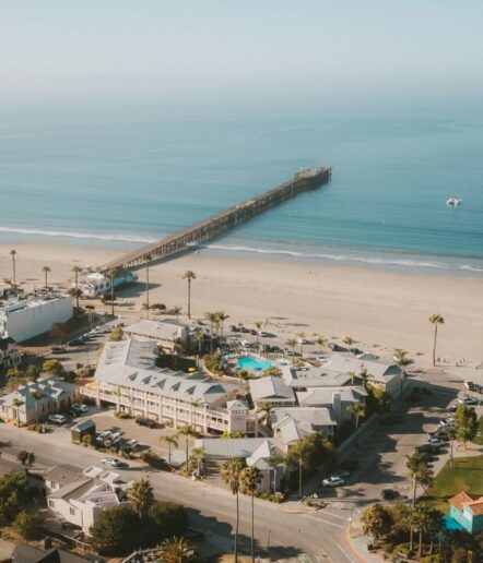 A long wooden pier extends over calm blue ocean water from a sandy beach, with coastal buildings, palm trees, and a swimming pool in the foreground. The sky is clear and the overall scene is peaceful.