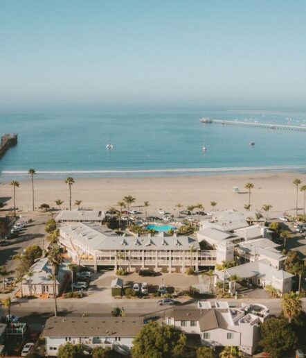 Aerial view of a beachside hotel complex, palm trees, sandy beach, and calm ocean with two piers extending into the water under a clear blue sky.