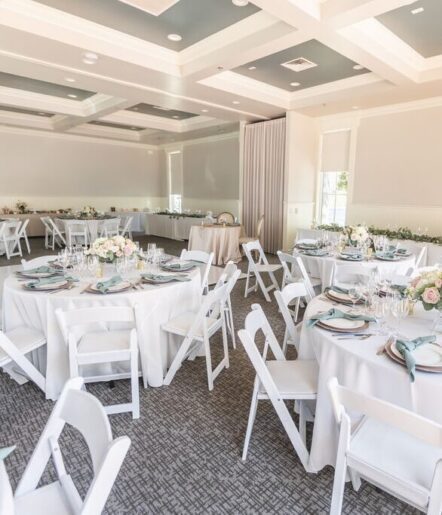 A decorated banquet hall with round tables covered in white tablecloths, set with plates, glasses, and blue napkins; white chairs surround the tables, and flower centerpieces add a soft touch.
