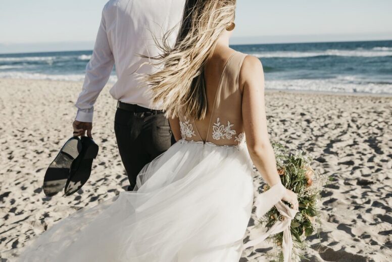 A bride and groom walk on a sandy beach. The bride holds flowers and wears a white gown, while the groom holds his shoes. The ocean and blue sky are in the background.