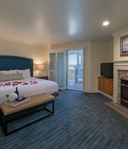 A cozy hotel room features a bed with red rose petals, a bench, a fireplace, a TV, and an outdoor balcony with seating. A tray with wine and flowers is on the bench; soft lighting creates a warm, inviting atmosphere.