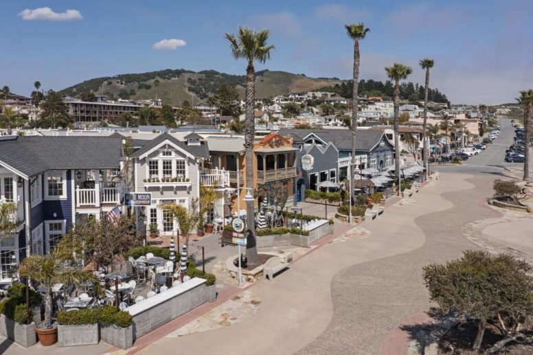 A coastal town street with colorful shops, outdoor dining areas, tall palm trees, parked cars, and green hills in the background under a blue sky with scattered clouds.