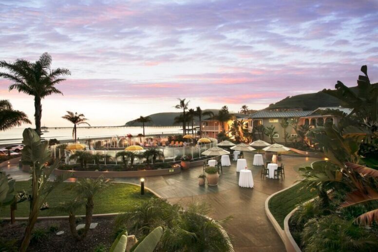 A coastal resort at sunset with palm trees, outdoor seating, cocktail tables, and a pool area overlooking the ocean. The sky is pink and purple with some clouds. Hills are visible in the background.