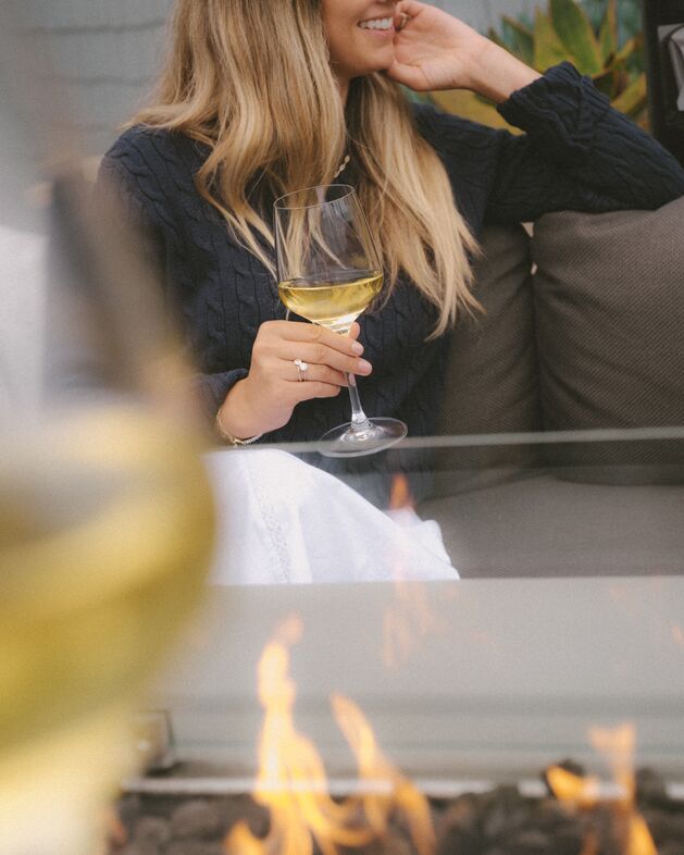 Woman with long blonde hair wearing a navy sweater sits on a couch, smiling and holding a glass of white wine. A fire pit burns in the foreground. Her face is partially out of frame.