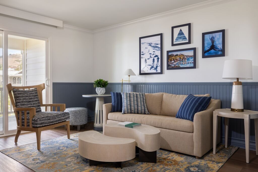 Modern living room with beige sofa, blue and striped pillows, patterned rug, wooden chair, abstract art on white walls, small round table, two modern coffee tables, lamp, and a sliding glass door letting in natural light.