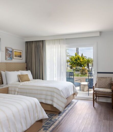 A bright hotel room with two double beds, a patterned rug, a chair, and large glass doors leading to a patio with outdoor seating and a view of palm trees and greenery.