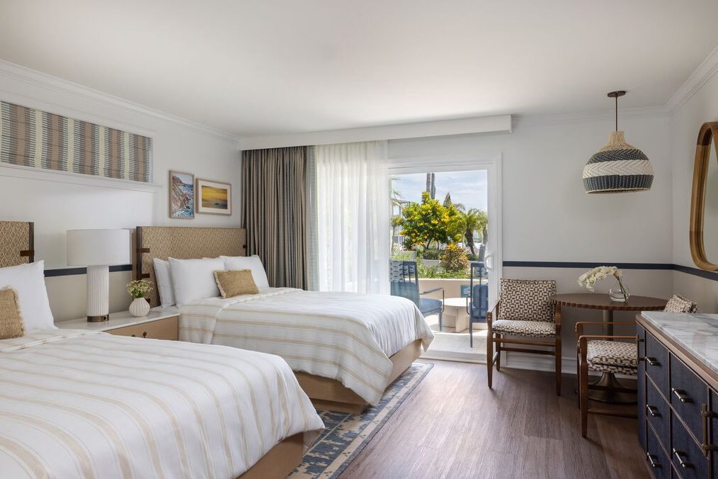 A bright hotel room with two double beds, a desk and chair, a dresser with a mirror, and a sliding glass door leading to a patio with outdoor seating and a view of trees and greenery.