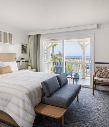 A bright hotel room with a large bed, a bench at the foot, a small sofa, and a balcony overlooking the ocean with chairs and palm trees visible outside.