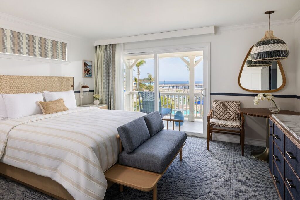 A bright hotel room with a large bed, a bench, a chair, a dresser with a vase, a mirror, and a balcony with an ocean and pier view through sliding glass doors.