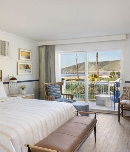 Bright hotel room with a bed, bench, chairs, and large window overlooking a balcony with outdoor seating, palm trees, and a view of hills and water. Decor includes coastal-themed artwork and neutral tones.