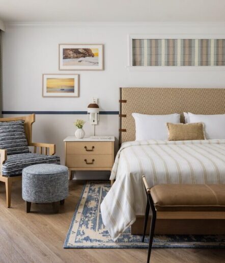 A cozy, modern bedroom with a bed, wooden nightstand, patterned chair, round ottoman, and artwork on the wall. Neutral tones and soft lighting create a warm, inviting atmosphere.