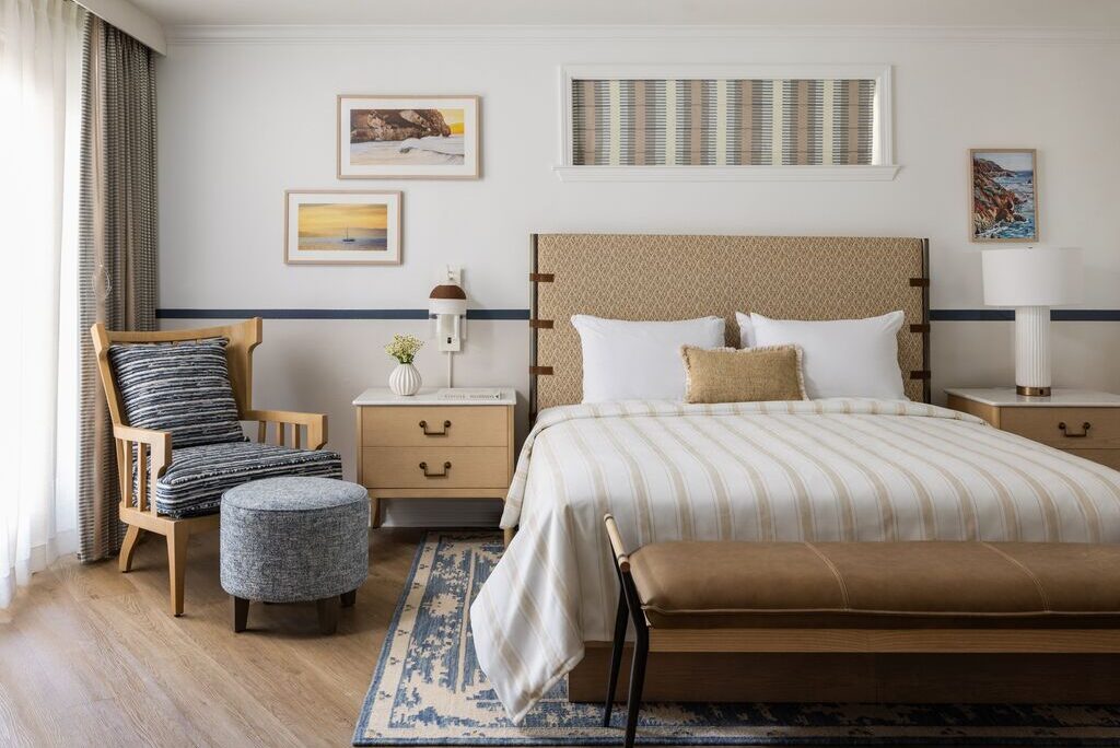A modern, cozy bedroom with a large bed, striped bedding, wooden bench, two nightstands, armchair with ottoman, wall art, and a patterned rug on a wood floor. Sunlight filters through sheer curtains.