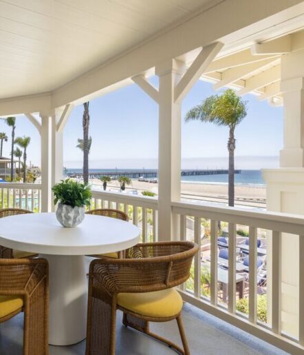 A round white table with four wicker chairs on a covered porch overlooks a beach, palm trees, and an ocean pier under a clear blue sky.