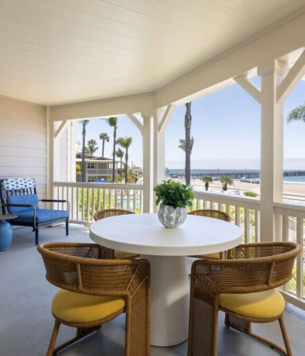 A covered porch with a round white table, four wicker chairs with yellow cushions, a potted plant centerpiece, and a view of palm trees and the ocean in the background.