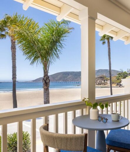 A balcony with two blue chairs and a small table overlooks a sandy beach, palm trees, mountains, and a long pier under a clear blue sky.