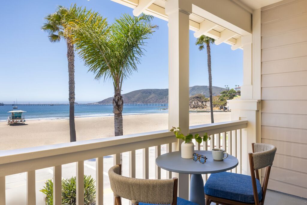 A small table with two chairs sits on a balcony overlooking a sandy beach, palm trees, a pier, and hills beneath a clear blue sky. A potted plant, book, and glasses rest on the table.