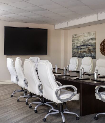 A modern conference room with a dark table, white chairs, water bottles, notepads, a large blank screen on the wall, and abstract artwork.