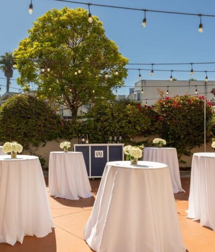 Outdoor event space with cocktail tables covered in white cloths, small flower centerpieces, string lights overhead, greenery, a tree, and a bar setup in the background under a clear blue sky.