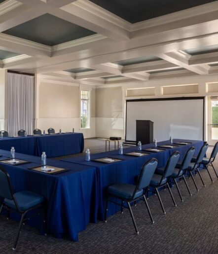 A conference room with U-shaped tables covered in blue cloths, arranged with chairs, notepads, and water bottles. There is a podium and a projection screen at the front.