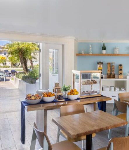 A bright cafe with wooden tables, a breakfast bar with fruit bowls, a pastry display case, and cereal dispensers. Glass doors open to an outdoor patio with palm trees.