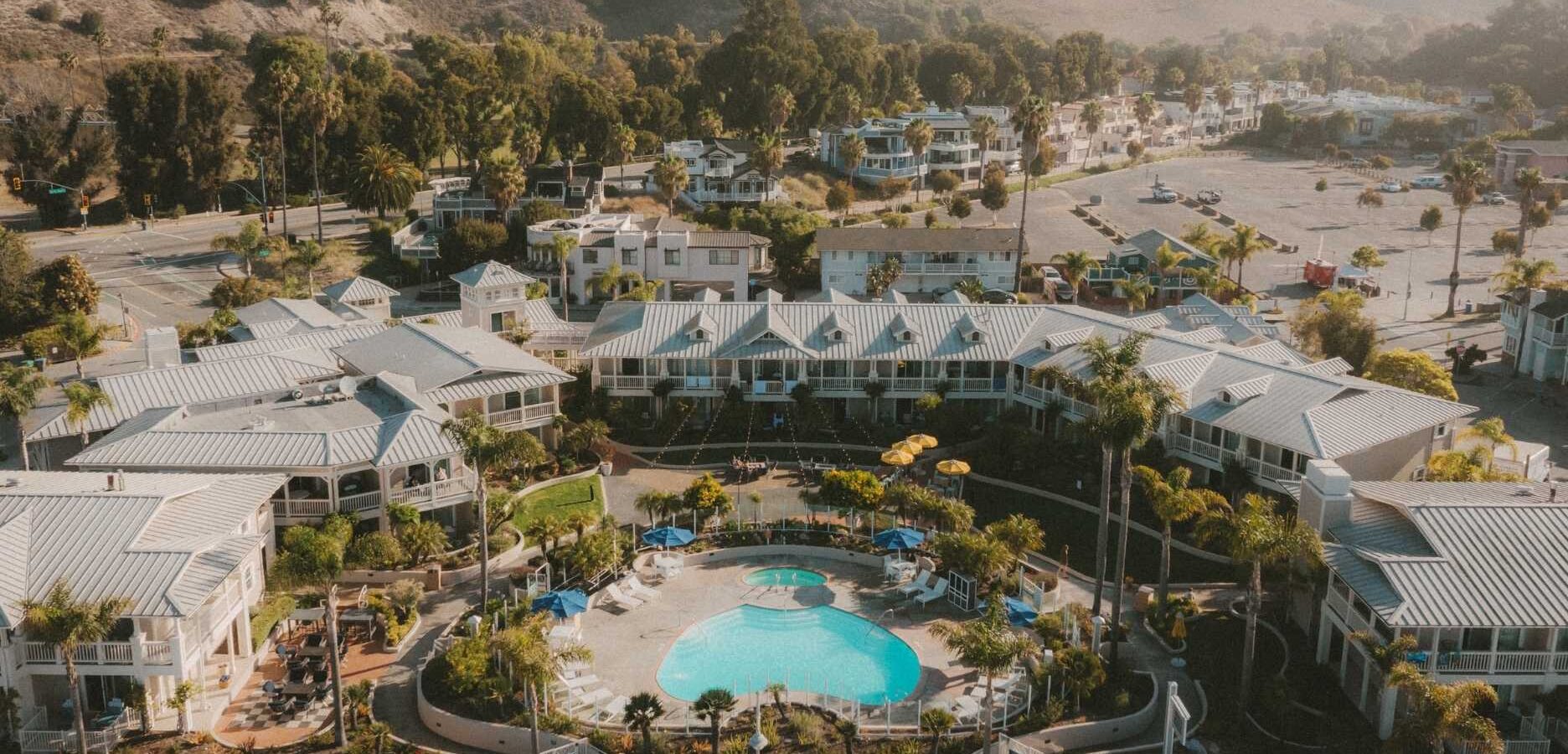 Aerial view of a resort with a central swimming pool, lounge chairs, umbrellas, and landscaped gardens, surrounded by buildings. Hills, trees, parked cars, and roads are visible in the background.