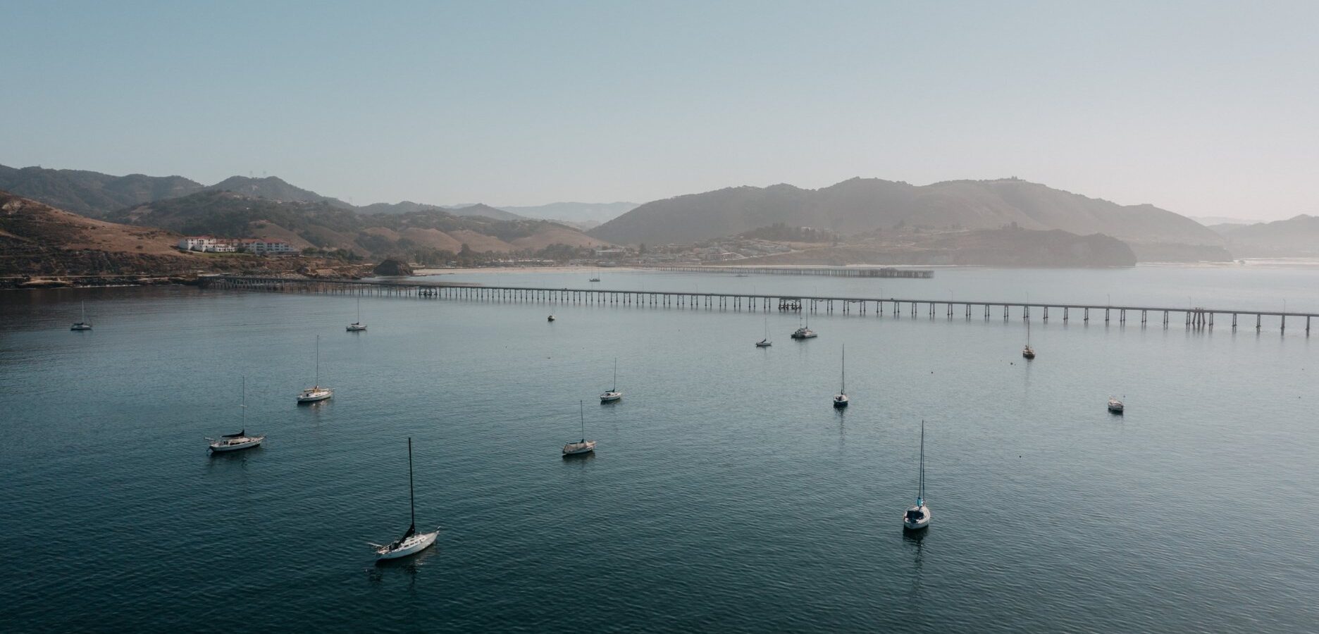 A calm bay with several sailboats anchored in the water, a long pier extending into the distance, and hills in the background under a clear sky.
