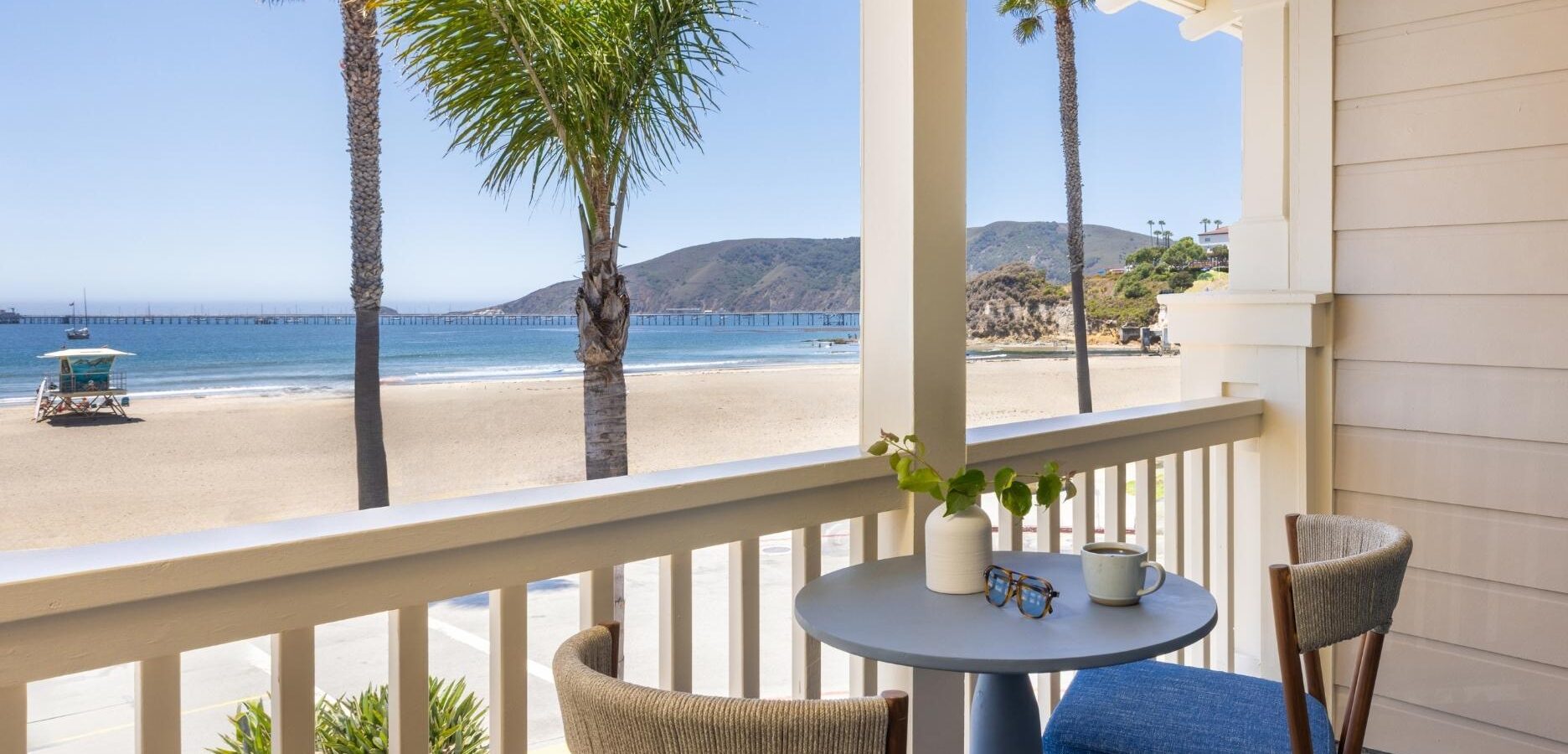 Cozy balcony with a small table and two chairs overlooking a sandy beach, palm trees, and the ocean with mountains and a pier in the distance on a sunny day.