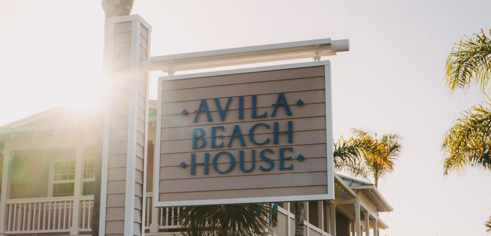 A wooden sign reading Avila Beach House in blue letters is shown with palm trees and a building in the background, sun shining brightly.