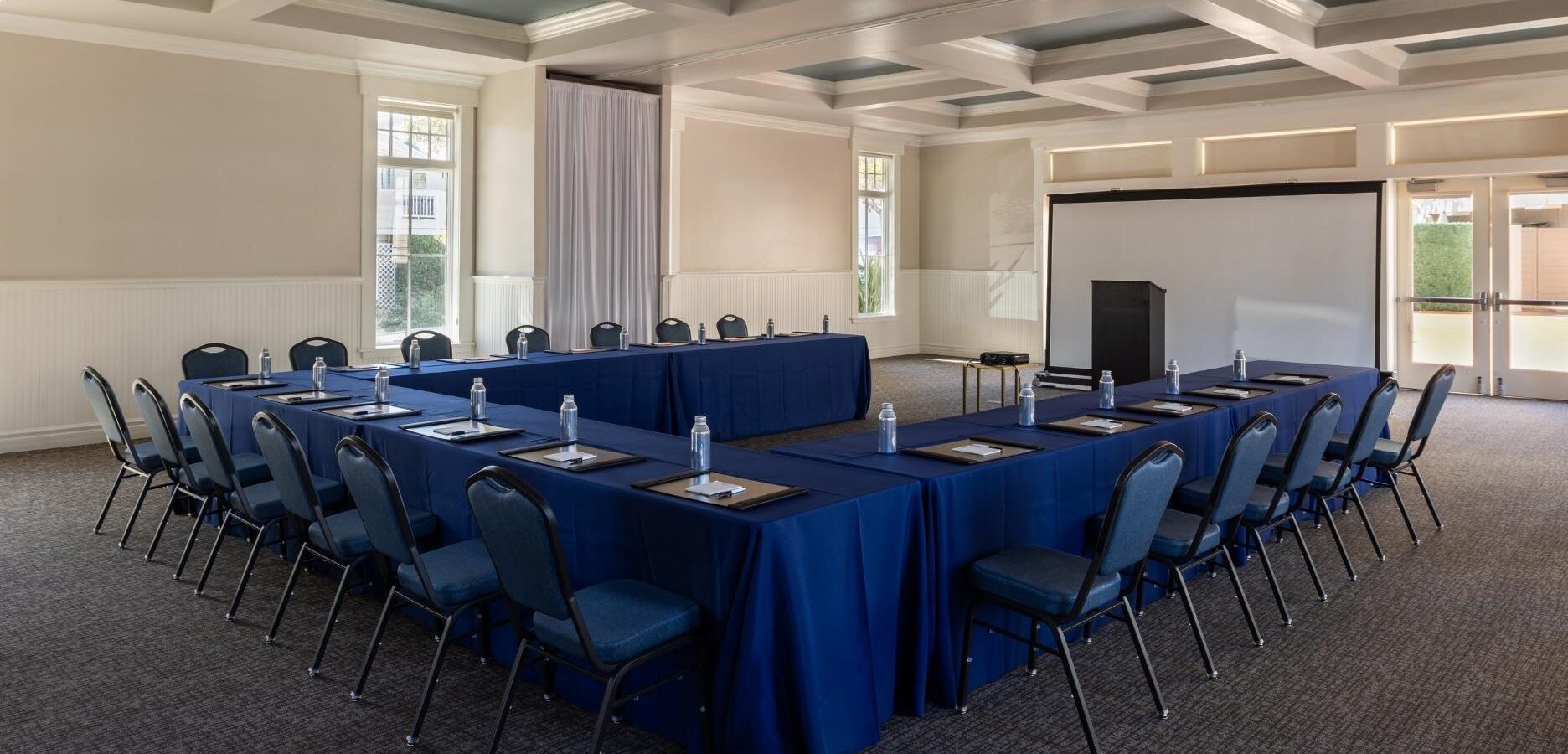 A conference room with U-shaped tables covered in blue cloth, each place set with a notepad, pen, and water bottle. Rows of blue chairs surround the tables, and a podium stands at the front near a projection screen.
