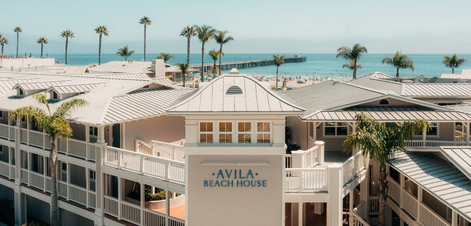 Avila Beach House with white railing, palm trees, and ocean in the background. A pier stretches into the water under a clear blue sky.