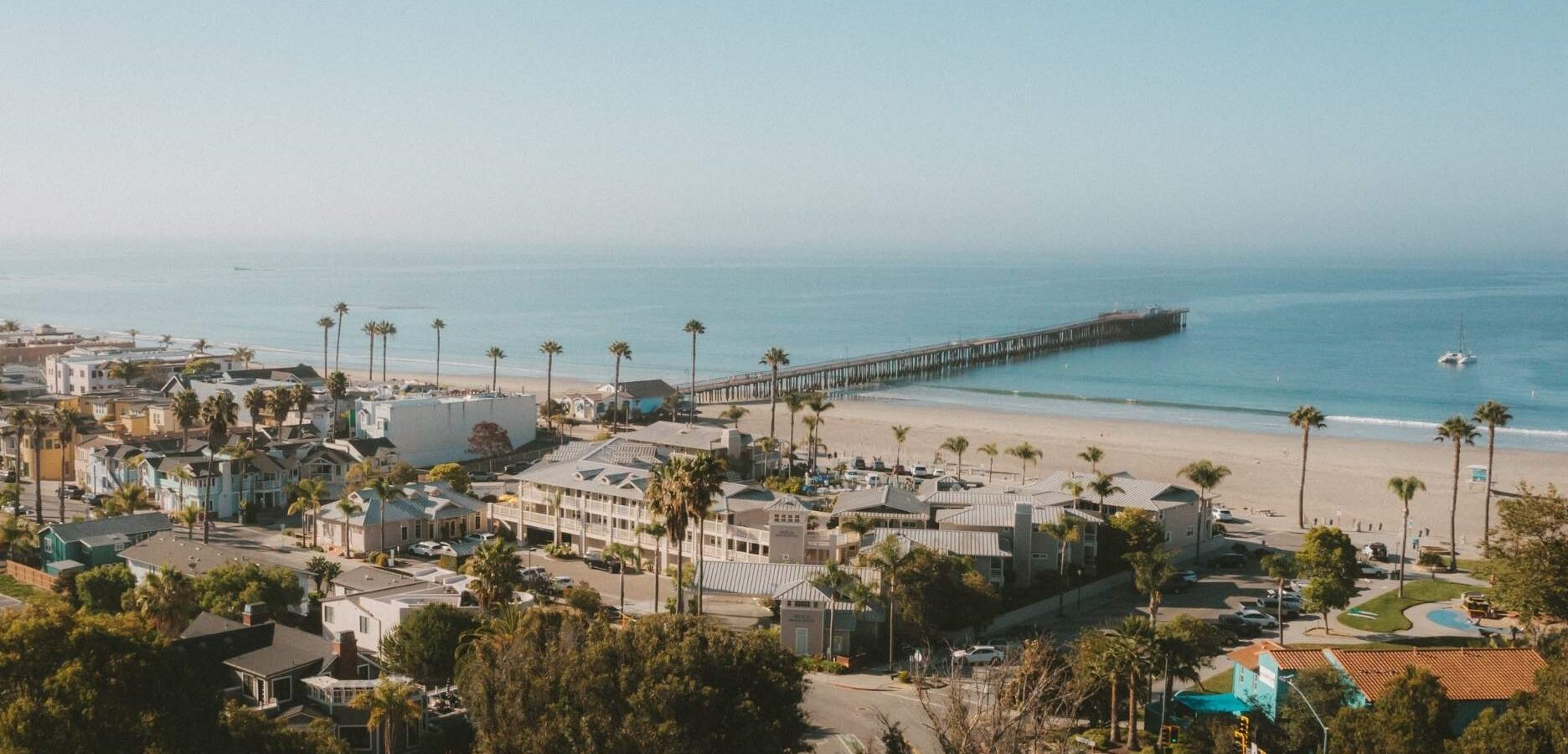 Aerial view of a beach town with palm trees, buildings, and a long pier extending into the calm blue ocean under a clear sky.