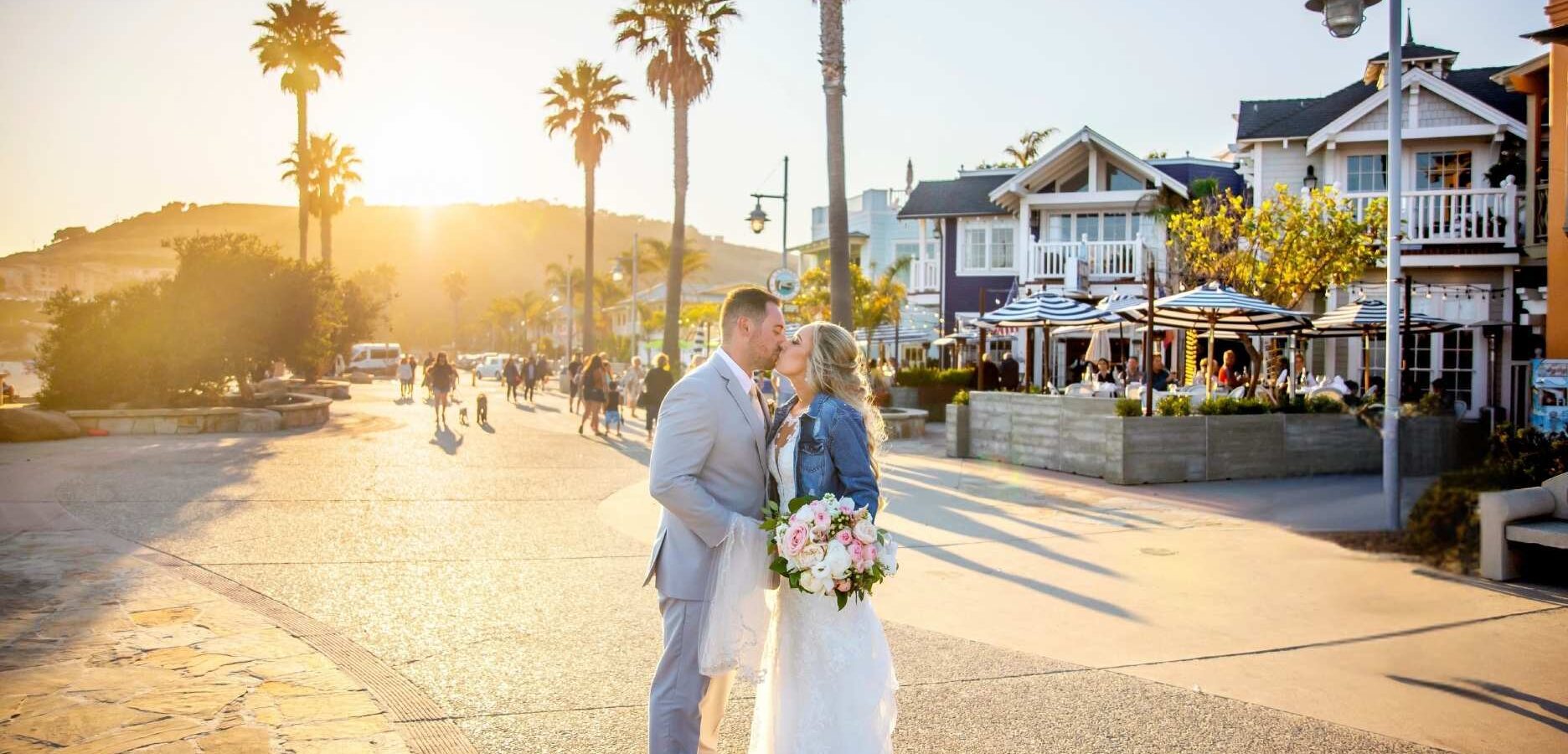 A bride and groom kiss on a sunny boardwalk lined with palm trees and beachside houses. The bride holds a bouquet and wears a jean jacket over her gown. People walk in the background.