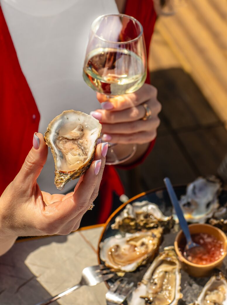 A person holds a glass of white wine and a freshly shucked oyster, with a plate of oysters and dipping sauce on the table below.