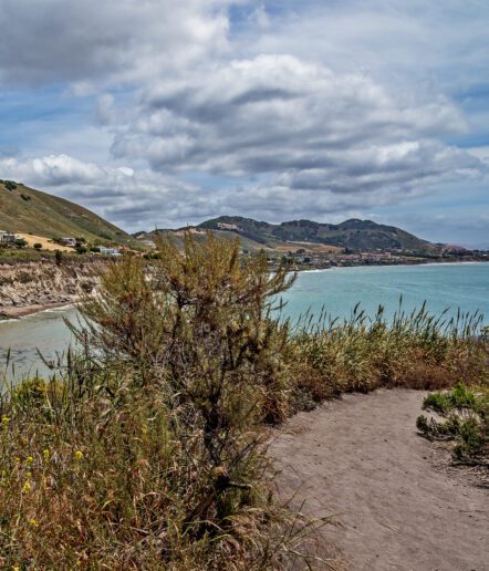 A dirt path lined with tall grass leads to a coastal cliff, overlooking turquoise water and distant hills under a partly cloudy sky.