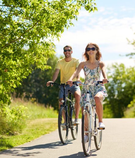 A man and a woman ride bicycles on a sunny, tree-lined path. Both are smiling and wearing sunglasses. The woman wears a summer dress, and the man wears a yellow shirt and shorts.